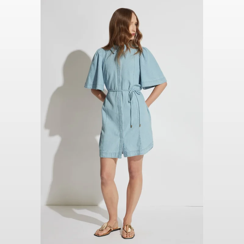Woman wearing a light blue denim Luna mini shirt dress with short flutter sleeves and a tie waist.