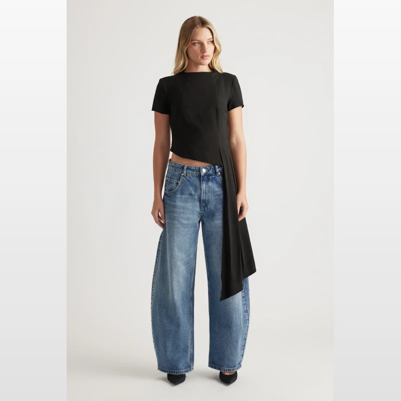 Woman wearing a black Sasha Drape Top with short sleeves, high neckline, and a side sash with pleat detail, paired with wide-leg blue jeans.