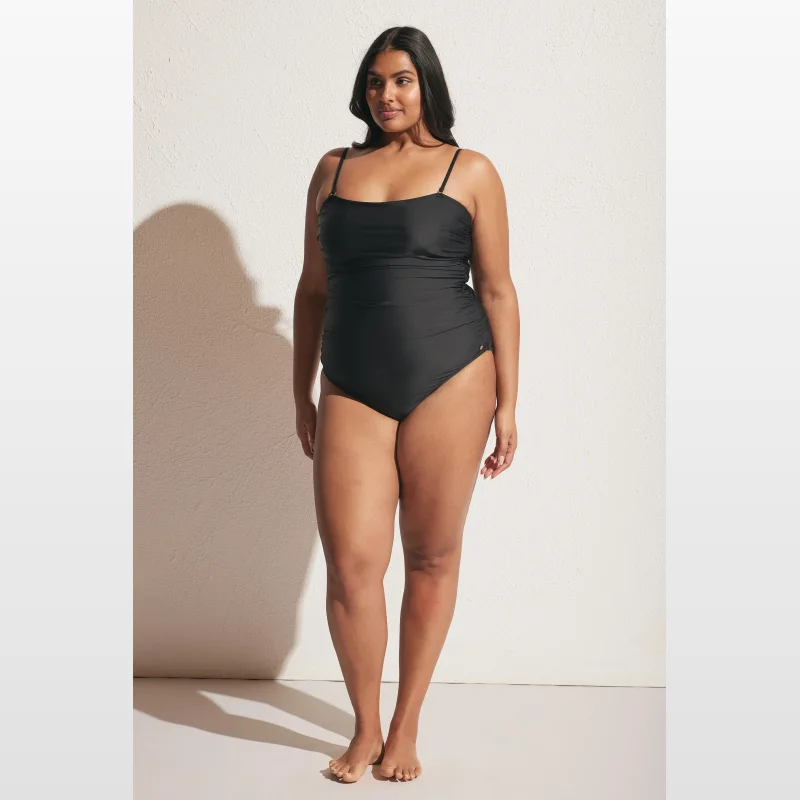 Woman wearing a black matte ruched one-piece swimsuit with a square neckline and thin adjustable straps.
