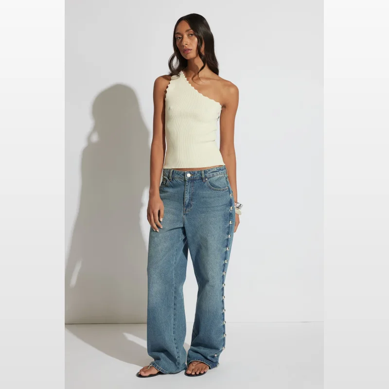 Woman wearing a cream ribbed one-shoulder tank top with an asymmetric scallop edge neckline and blue wide-leg jeans.
