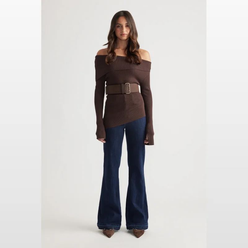 Woman wearing a brown ribbed off-shoulder knit top with long sleeves and an asymmetrical hem, paired with dark wash flared jeans and a brown belt.