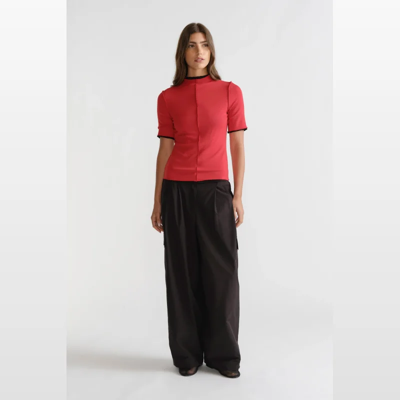 Woman wearing a red D-Luxe short sleeve mock neck top with black trim and centre front seam detail, paired with wide-leg black trousers.