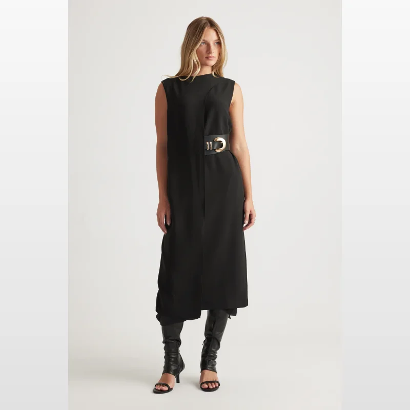 Woman wearing a black sleeveless midi dress with a high round neckline, an attached belted waist detail with a gold buckle, and a back split. She is also wearing black knee-high heeled boots.