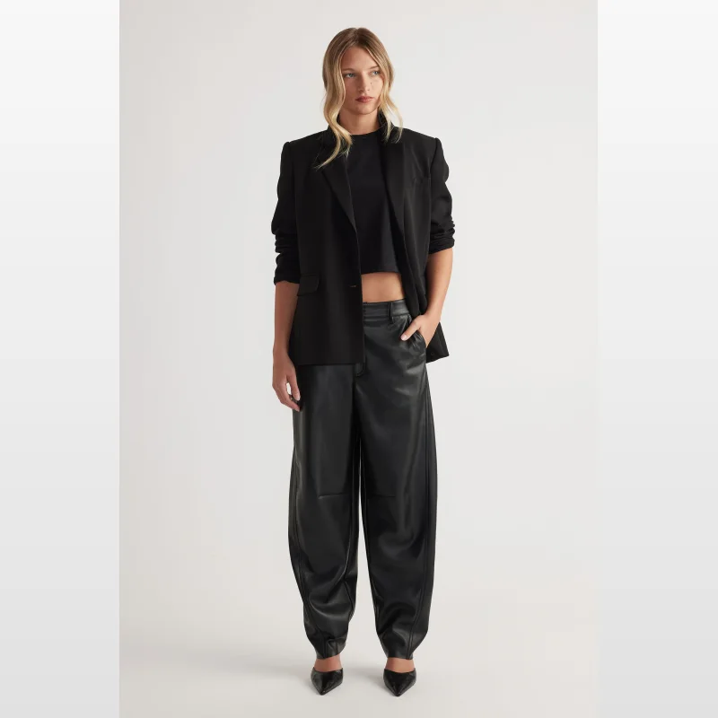 Woman wearing Decjuba Natalie Faux Leather Barrel Leg Pants in black, paired with a black crop top and black blazer.