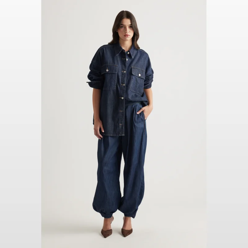 Woman wearing a dark wash denim shirt with collared neckline, full length sleeves with deep cuff detail, and two functional chest pockets. The shirt has a button-through front and a curved hemline.
