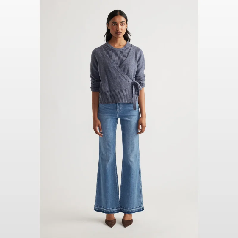 Woman wearing a blue wrap knit jumper with a built-in tank top and tie detail at the side, paired with flared denim jeans.