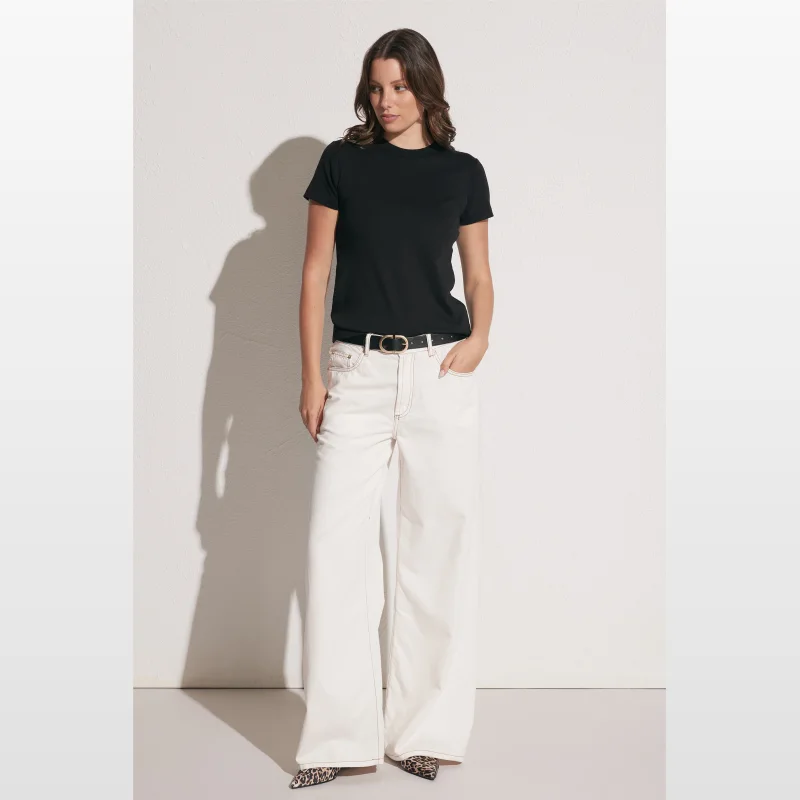 Woman wearing a black crew neck t-shirt and white wide-leg jeans, standing against a white textured wall.