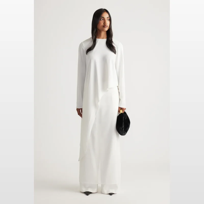 Model wearing a white Codi Satin Drape Top with long sleeves and an asymmetric hem, paired with white wide-leg pants.