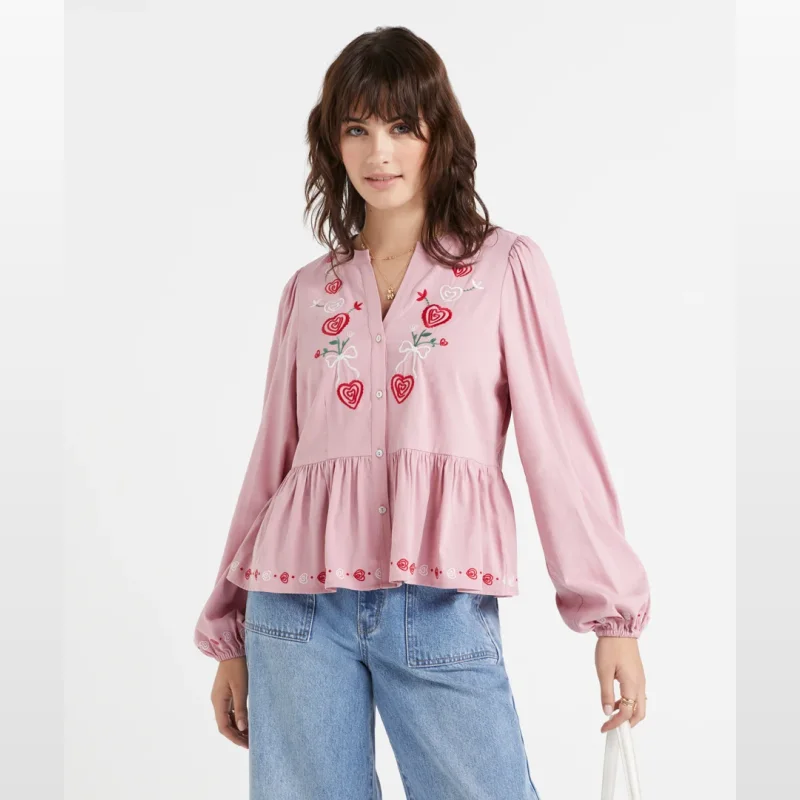 Woman wearing a pink long-sleeved blouse with heart embroidery on the front and hem, paired with blue jeans.