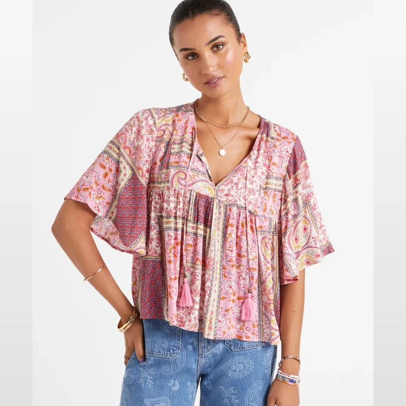 Woman wearing a pink and multi-coloured patchwork print boho blouse with short sleeves and tassel details on the front.