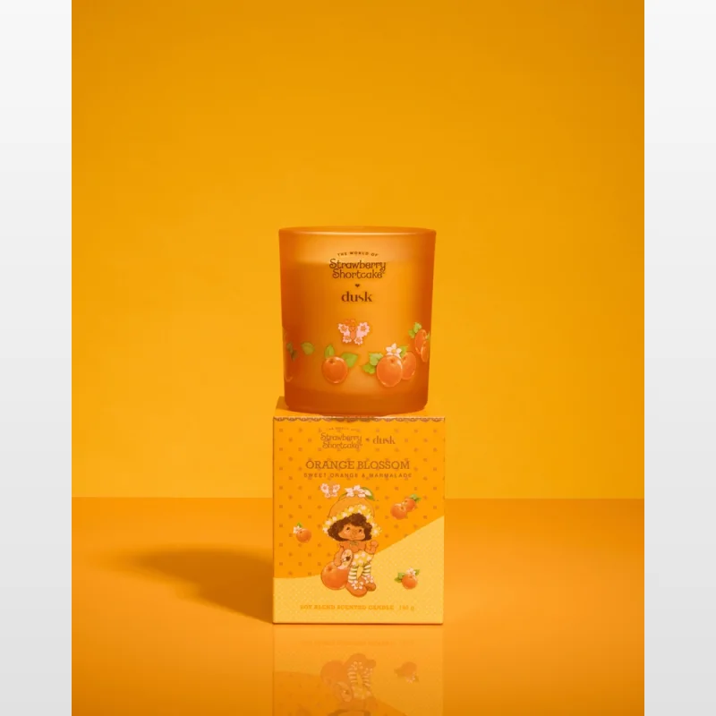 A Dusk Orange Blossom scented candle in a frosted orange glass jar, with a matching box featuring Strawberry Shortcake artwork. The candle and box are displayed against a vibrant orange background.