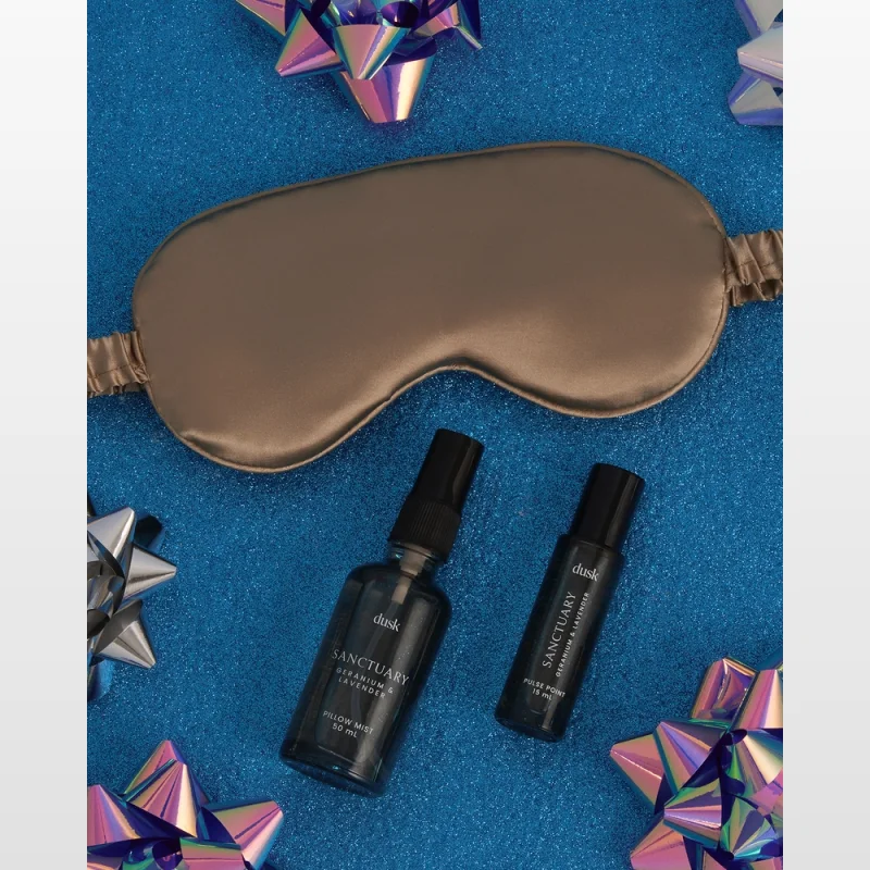 A taupe satin eye mask rests on a sparkly blue surface, accompanied by a bottle of Dusk Sanctuary Geranium & Lavender Pillow Mist and a Dusk Sanctuary Geranium & Lavender Pulse Point rollerball, surrounded by iridescent gift bows.