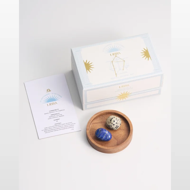 A Libra zodiac crystal gift box from Dusk, containing a small wooden dish with a blue lapis lazuli stone and a spotted dalmatian jasper stone, alongside an information card.