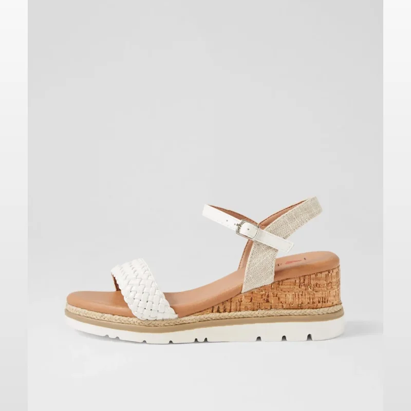 Side view of a white Fleurie sandal with a woven toe strap, a buckled ankle strap, and a cork-style wedge heel.