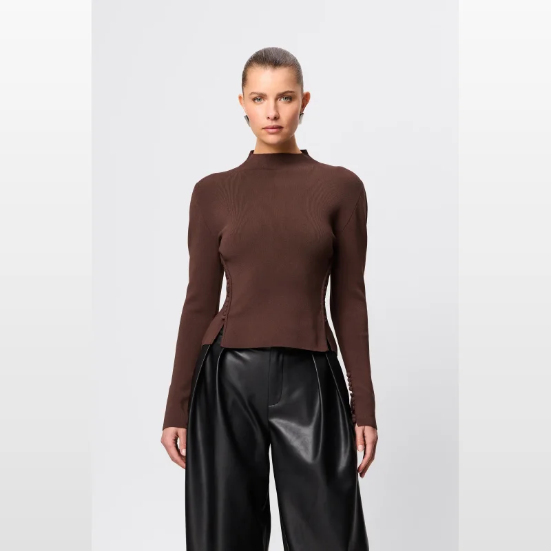 Woman wearing a brown long-sleeved knit top with a mock neckline and button details on the sleeves and sides, paired with black leather pants.
